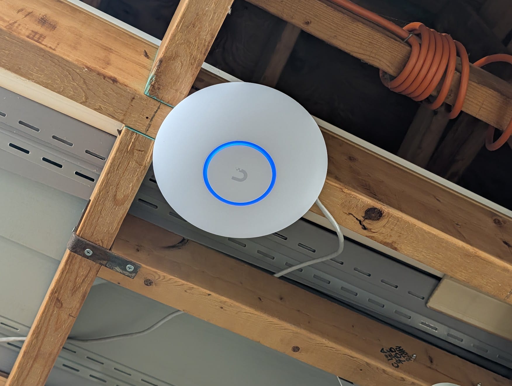 A UniFi U7 Lite access point mounted to basement ceiling joists with a blue LED ring