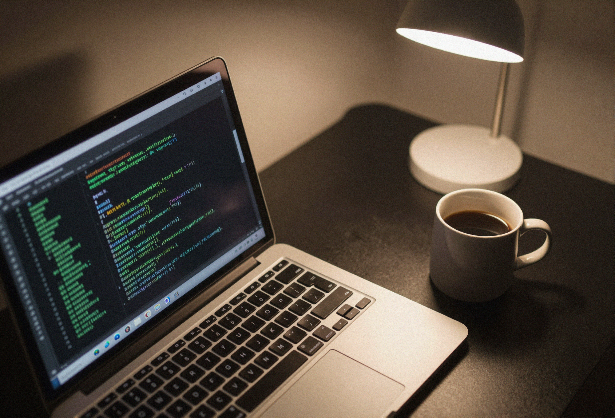 A laptop on a dark desk showing code in a terminal, lit by a single desk lamp, with a mug of coffee beside it