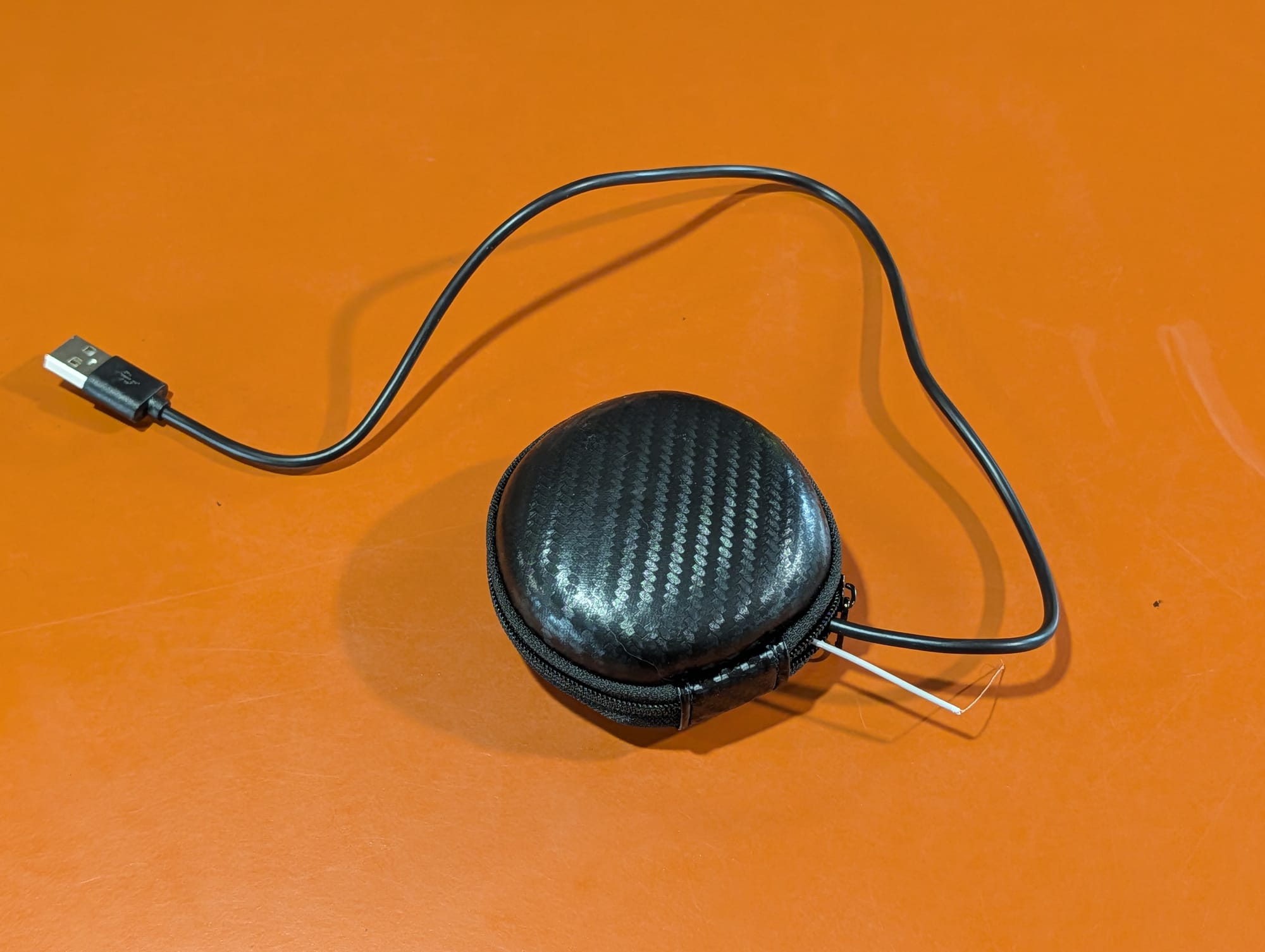 A carbon-fiber zippered earbud case, closed, with a USB cable coming out of it